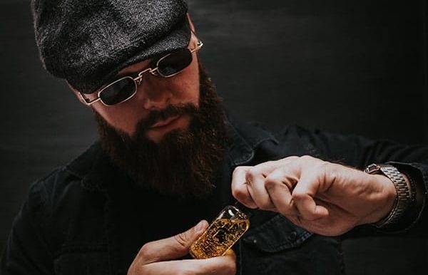 Čovek sa bradom i naočarima u crnoj košulji otvara Warrior Pure Gold bočicu sa uljem za bradu.